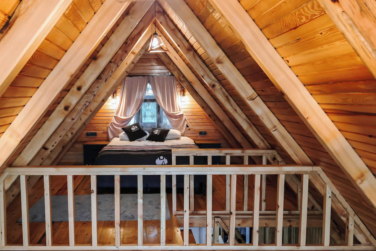 A high-ceilinged attic bedroom with a wooden sloped roof, a bed with two pillows, and a view of the outside through a window.