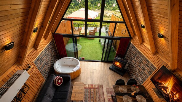 Un acogedor dormitorio en el ático con un sofá negro, una mesa de madera con individuales tejidos, una chimenea y un gran ventanal con vistas a un césped verde.