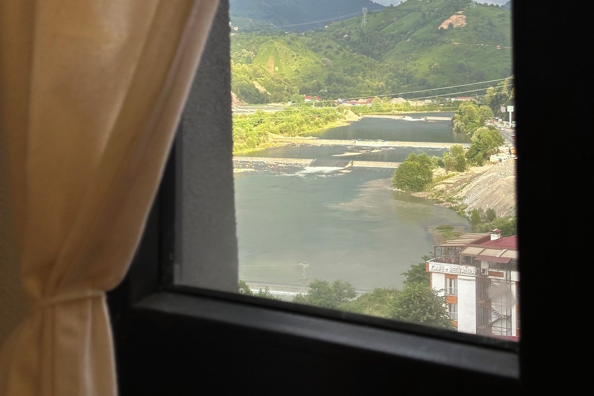 Vista pela janela com cortinas, mostrando um rio, colinas verdes e um edifício com a placa 'CAFE RESTAURANT'.