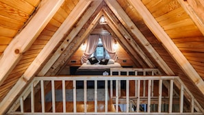 Un dormitorio en el ático con techos altos, techo inclinado de madera, una cama con dos almohadas y vistas al exterior a través de una ventana.