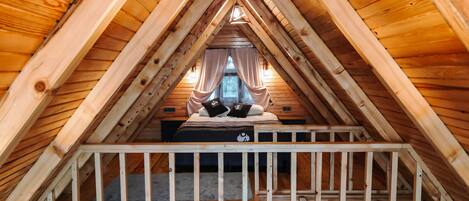 A high-ceilinged attic bedroom with a wooden sloped roof, a bed with two pillows, and a view of the outside through a window.