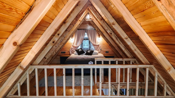 A triangular attic bedroom with wooden beams, a bed with two pillows, and a window with curtains.