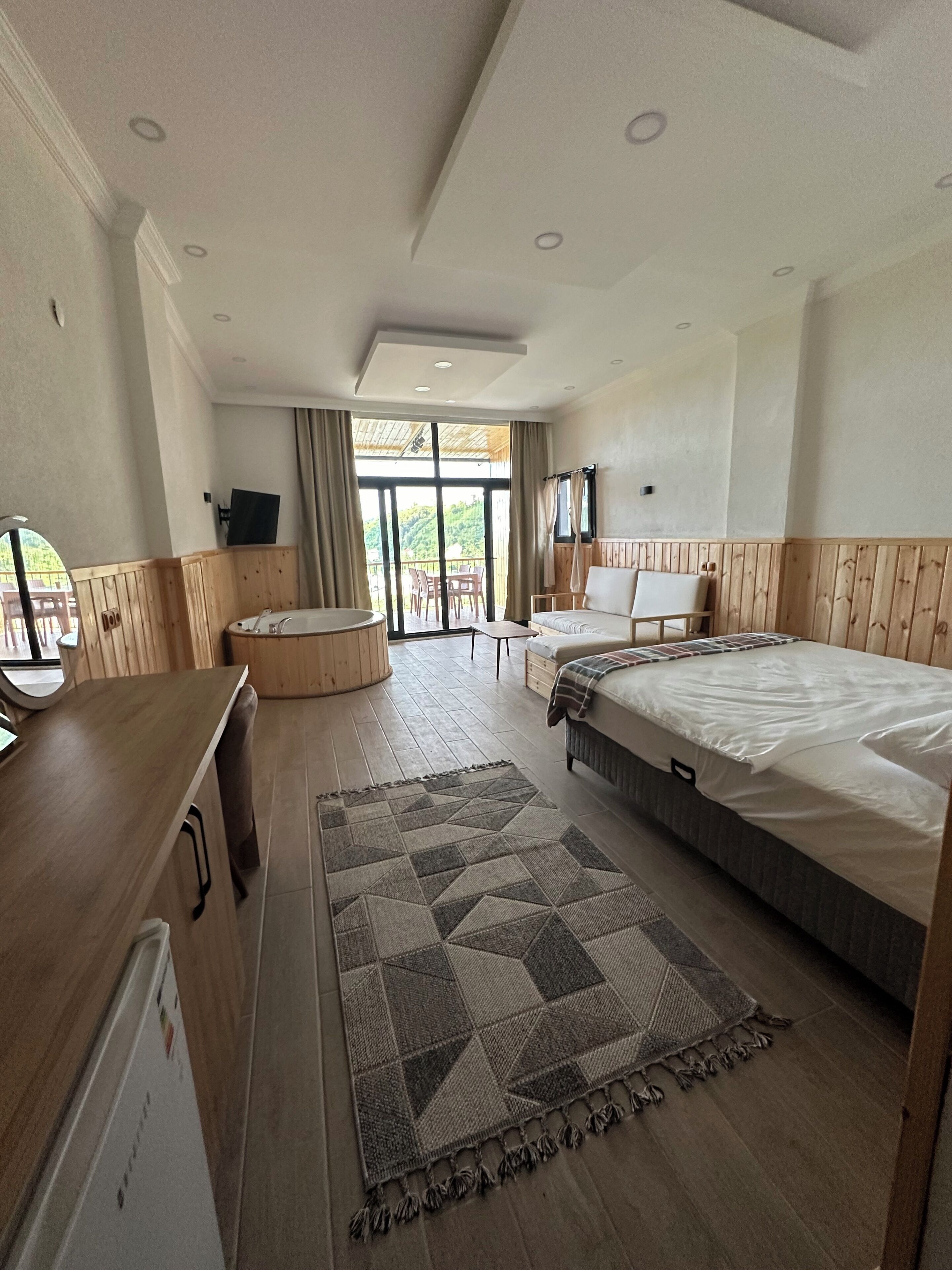 A hotel room with a bed, a desk, a TV, a balcony with a table and chairs, and a large window.