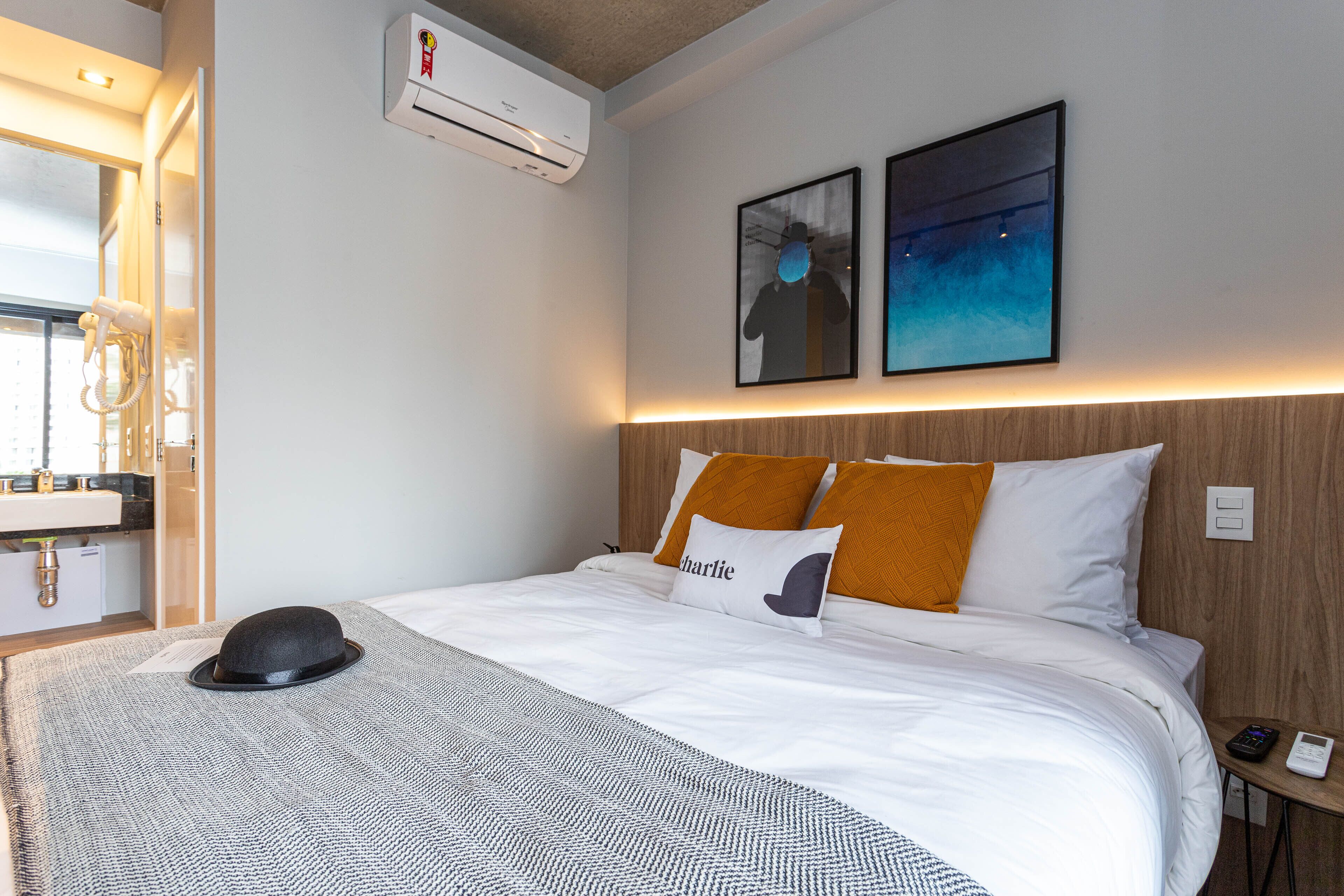 A hotel room with a bed, a hat on the bed, a nightstand with a phone, and a wall-mounted air conditioner.