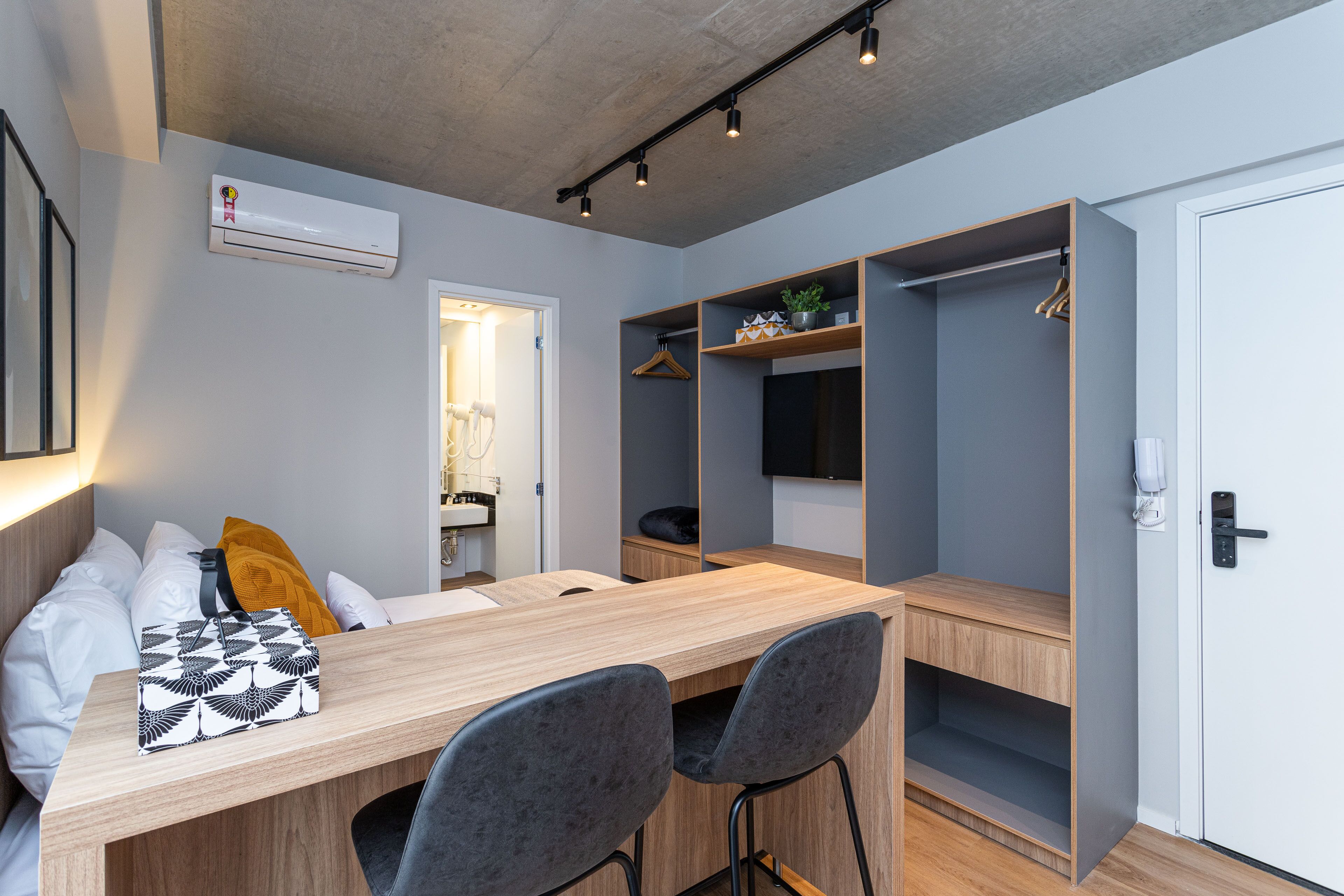 Una habitación moderna con una mesa de comedor de madera, dos sillas grises, una cama con almohadas blancas y amarillas, un armario y un televisor montado en la pared.