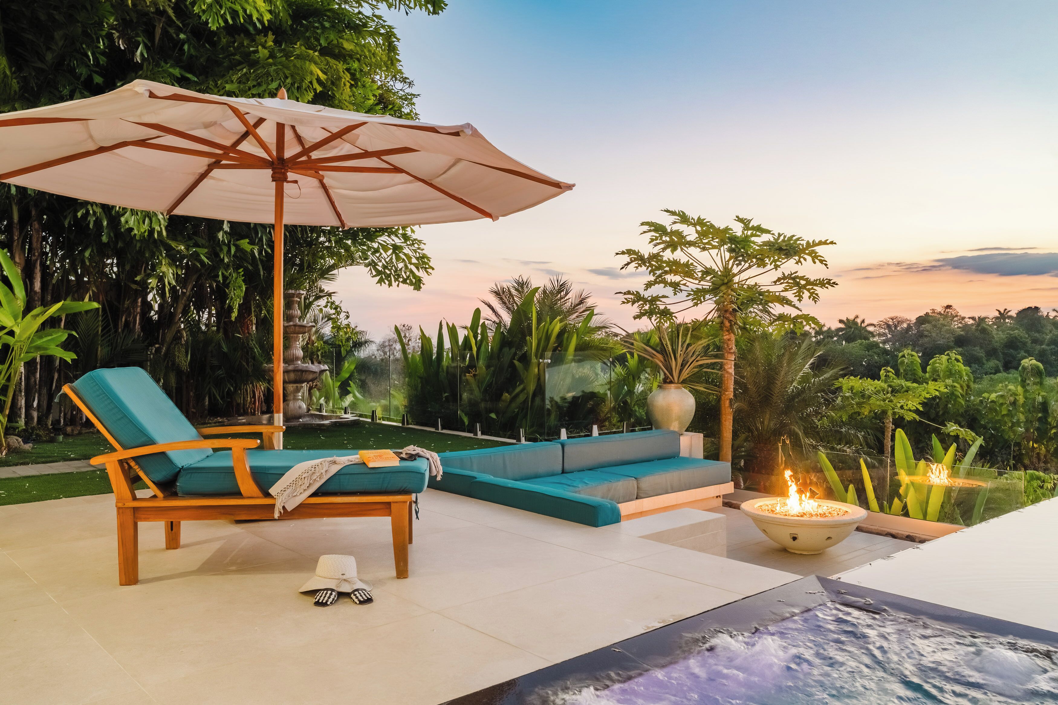 A lounge area with a wooden chair, a sofa, an umbrella, and a fire pit.