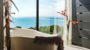 A bathroom with a bathtub, a window with a view of the sea, and a plant.