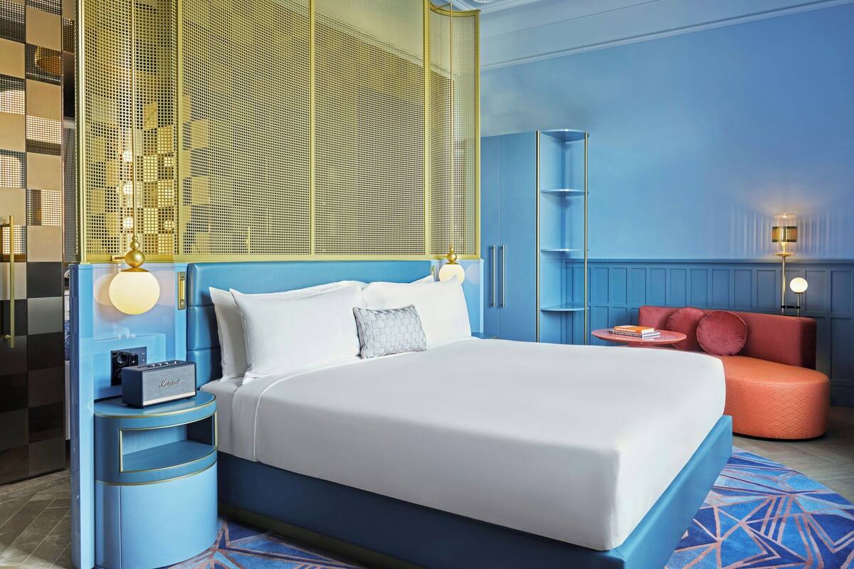A modern hotel room with a large bed, a nightstand with a lamp, a red sofa, and a decorative wall panel.