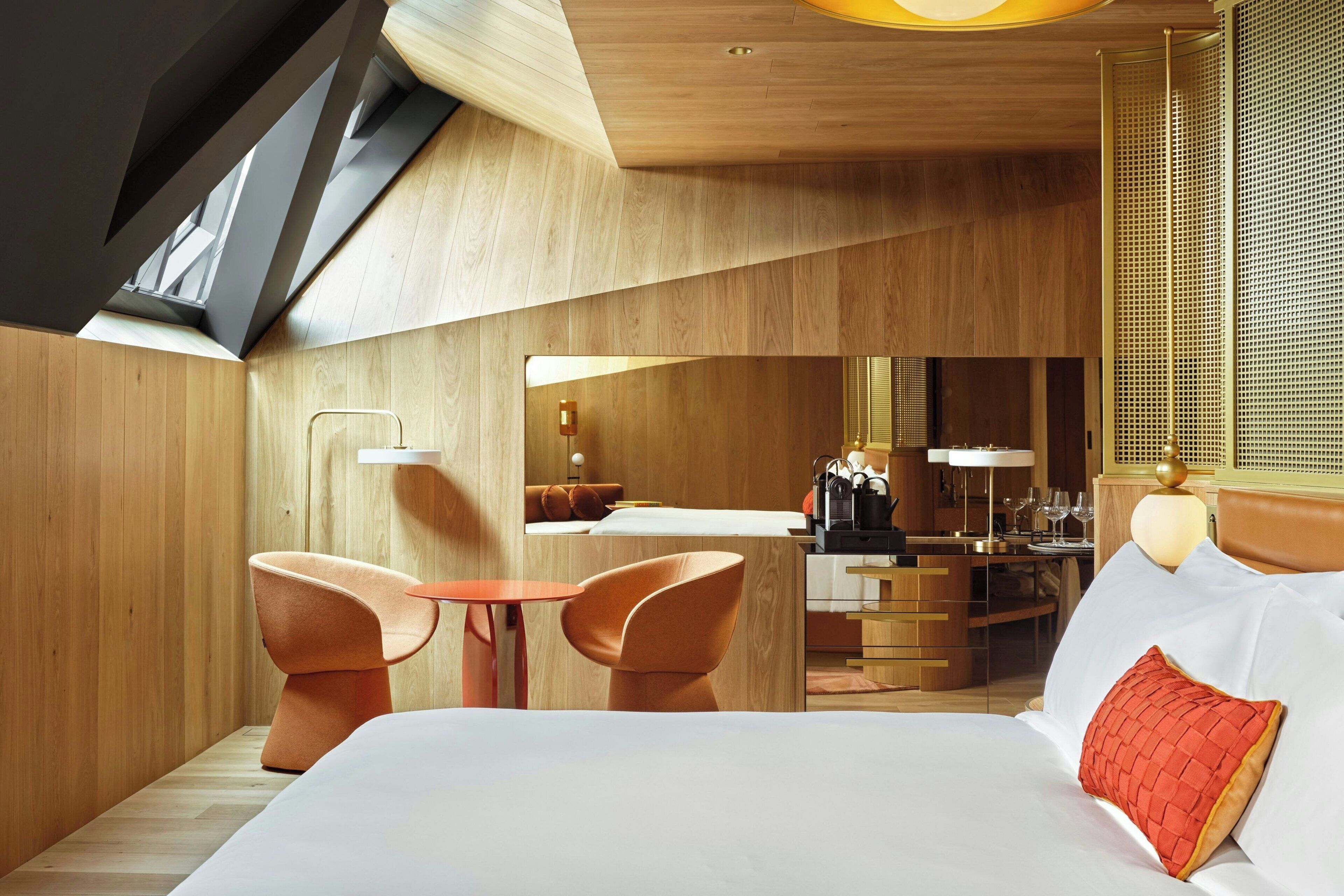 A modern hotel room with a wooden interior, a bed with white linens, a red accent pillow, and a seating area with two orange chairs.