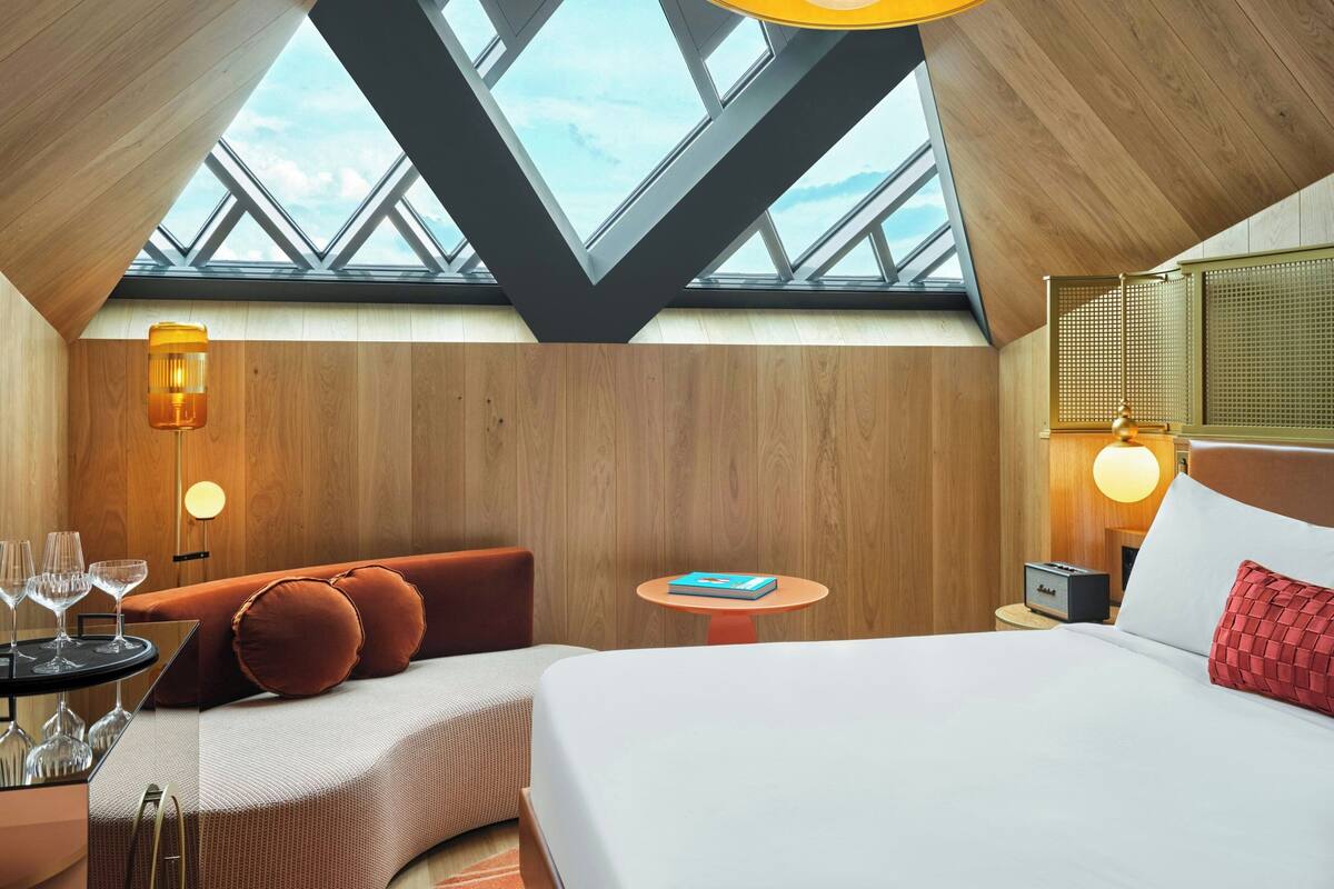 A modern hotel room with a skylight, wooden walls, a bed, a sofa, a small table with a book, and a nightstand with a lamp.