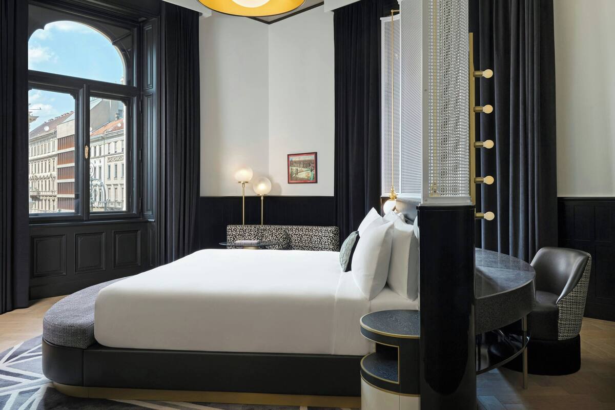 A modern hotel room with a large bed, a round table, and a chair. There is a window with a view of buildings outside.