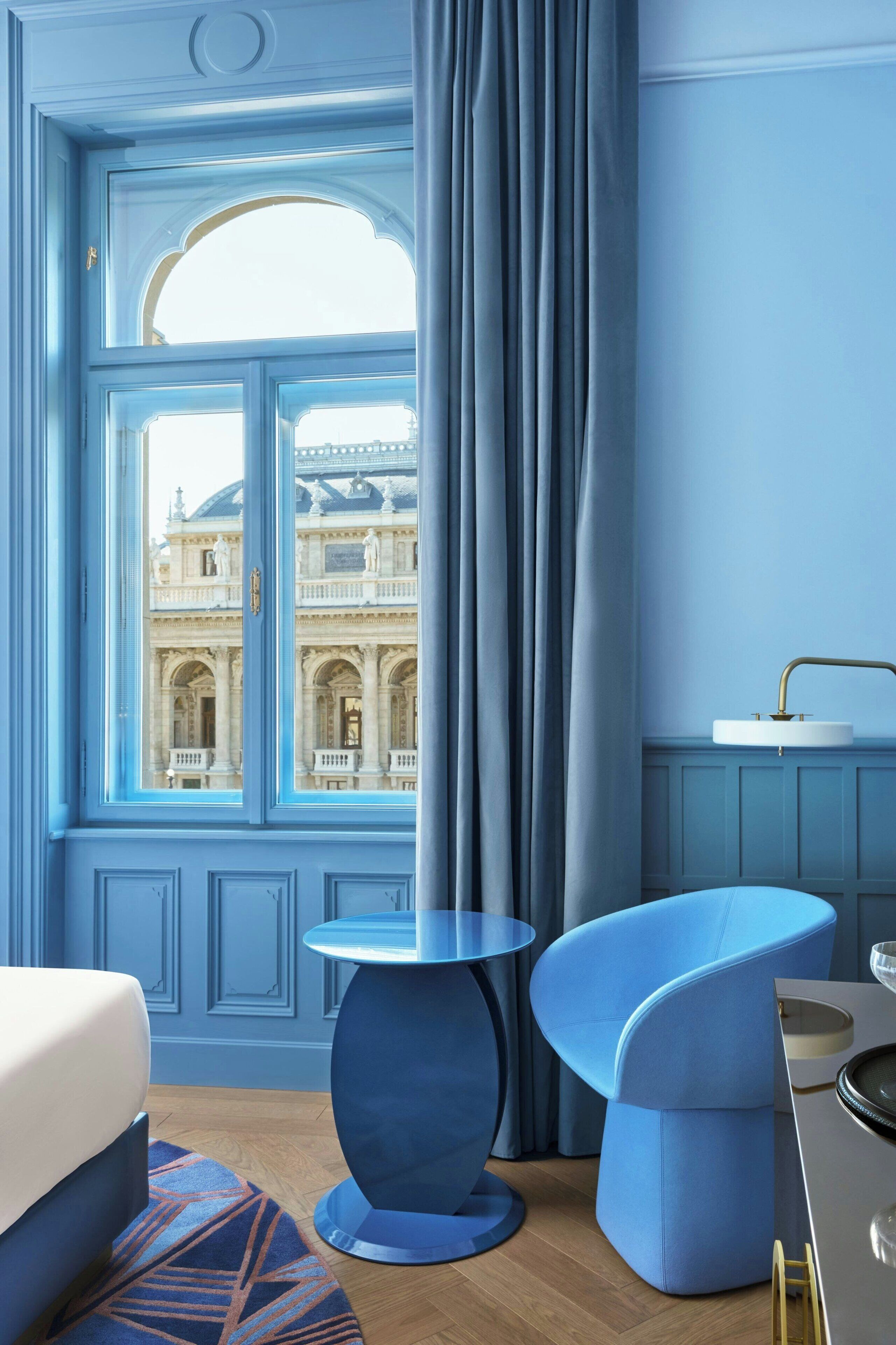 Una habitación con un ventanal grande, una cama, una mesa redonda y una silla azul.