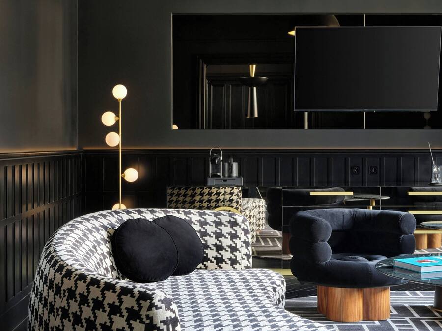 Una sala moderna con un sofá estampado de pied de poule, un sillón oscuro y una mesa redonda de centro. Un televisor de pantalla plana grande está montado en la pared, y hay una lámpara de pie con varias bombillas.