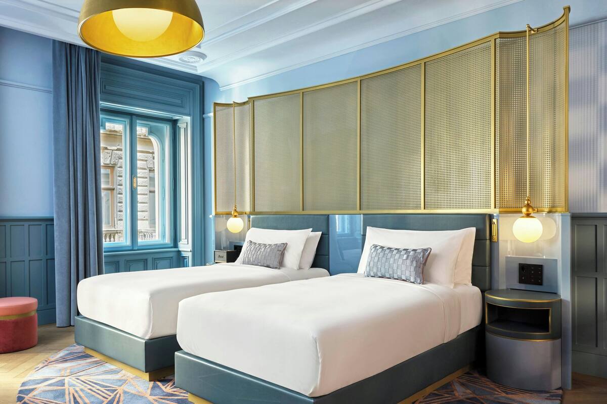 A hotel room with two beds, a large window, a decorative wall panel, and a nightstand with a lamp.