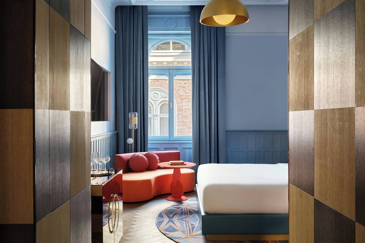 A modern hotel room with a bed, a red sofa, a small table, and a view of a building through a window.