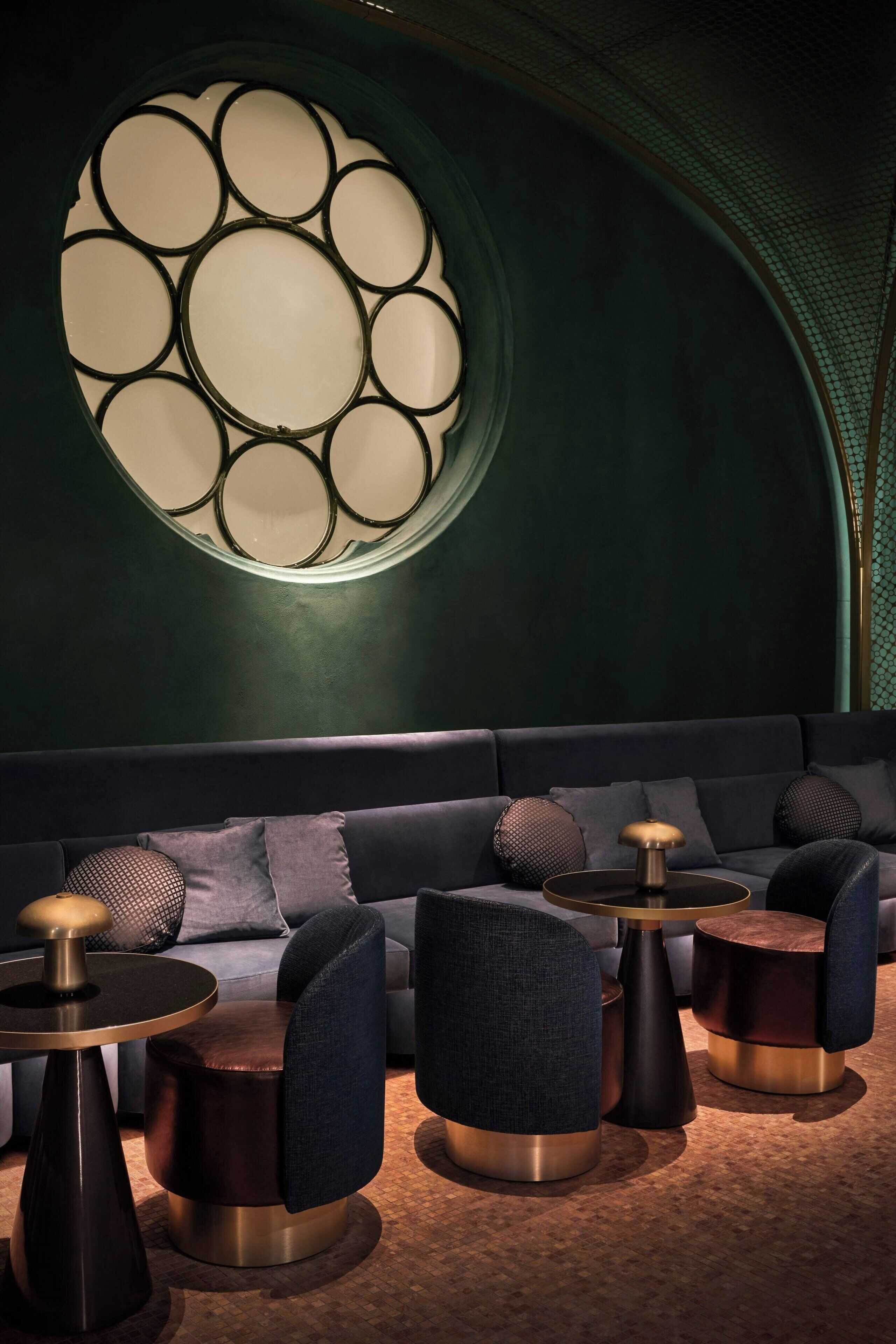 Ein stilvoller Lounge-Bereich mit einer dunkelgrünen Wand, einer runden Leuchte und einem dunkelblauen Sofa mit passenden Sesseln und runden Tischen.