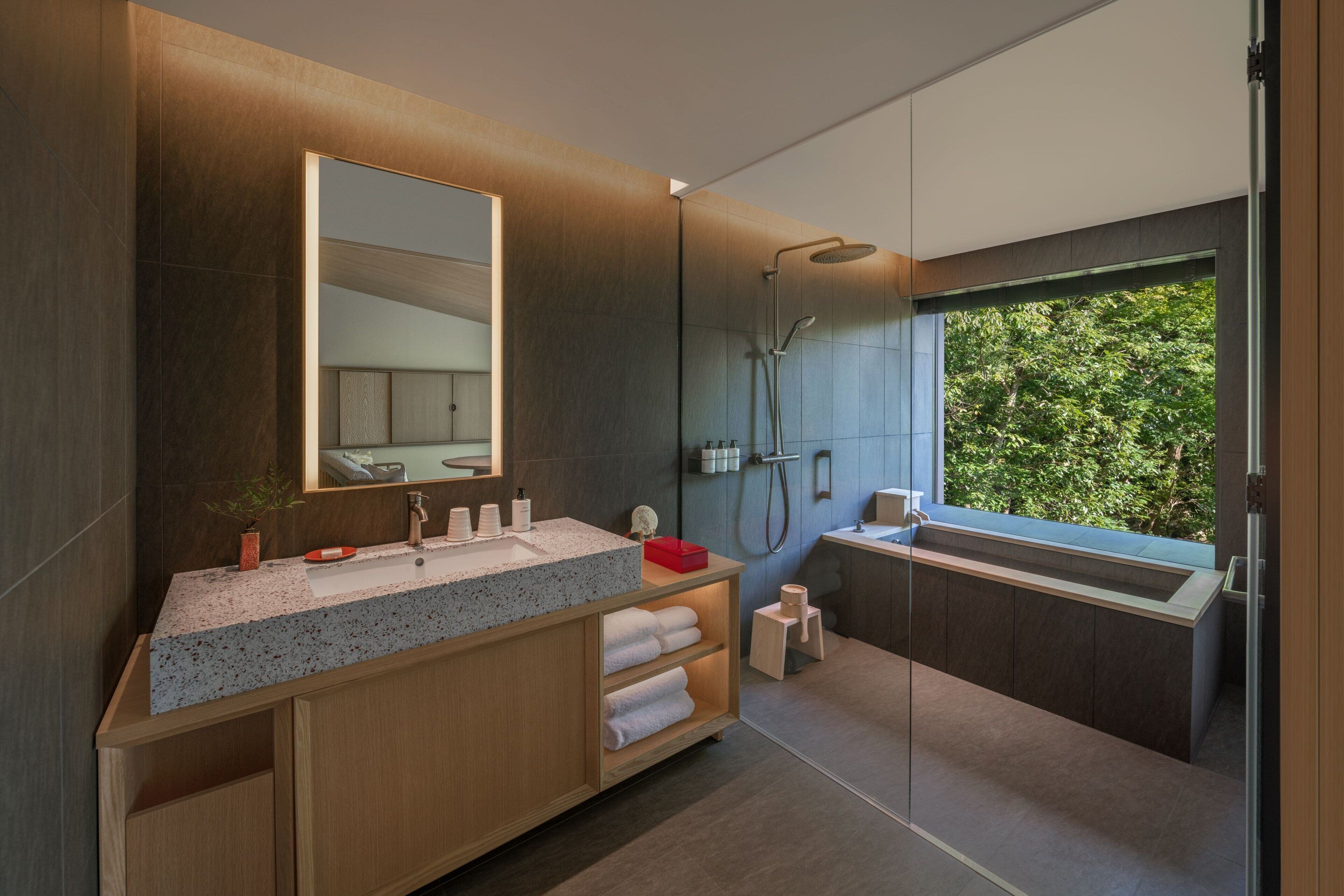 A modern bathroom with a large mirror, a shower, and a window with a view of greenery.