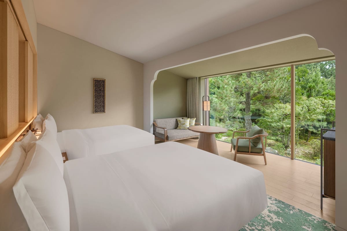 Habitación de hotel con dos camas, un sofá, una mesita y un balcón con vista a árboles.