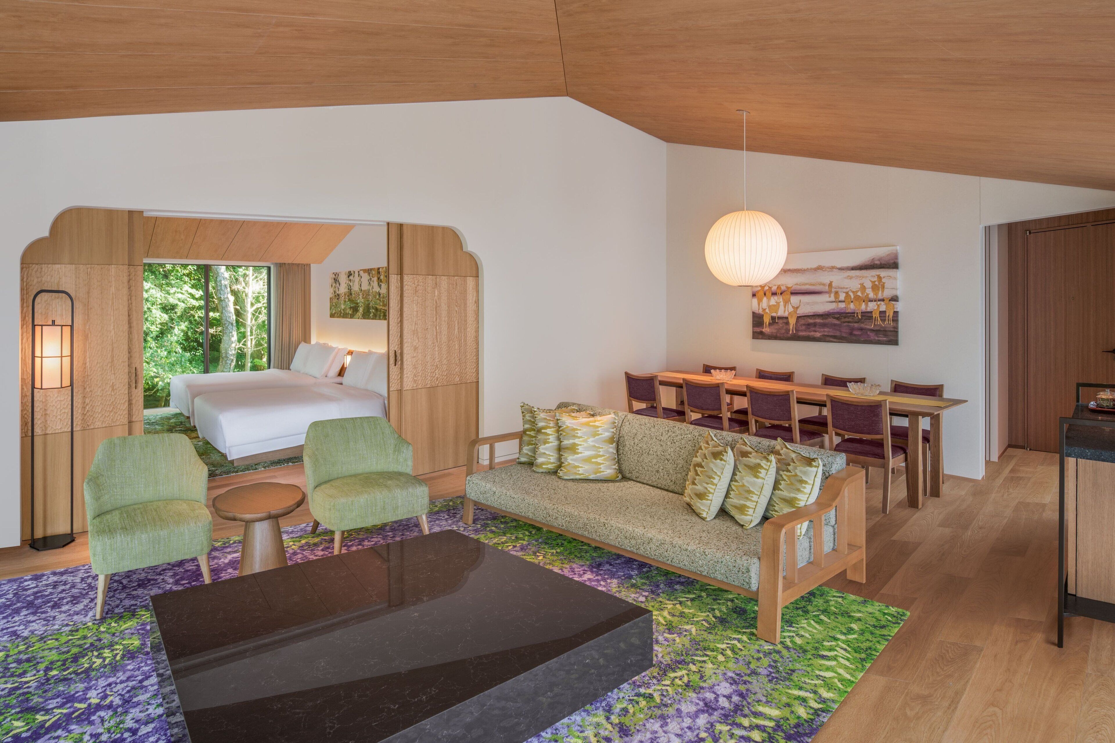 A modern hotel room with a bed, green armchairs, a sofa, a dining area, and a large window.
