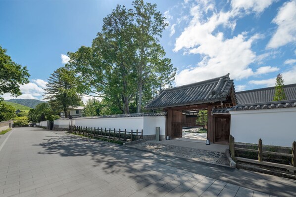 A traditional Japanese gate with a wooden structure and tiled roof, leading to a pathway lined with white walls and a wooden fence.