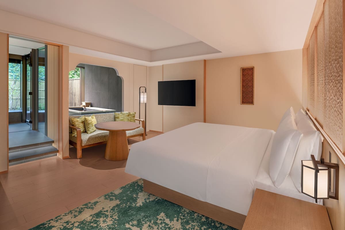 Una habitación de hotel moderna con una cama grande, una mesita, un sofá, un televisor y un baño visible a través de una puerta abierta.