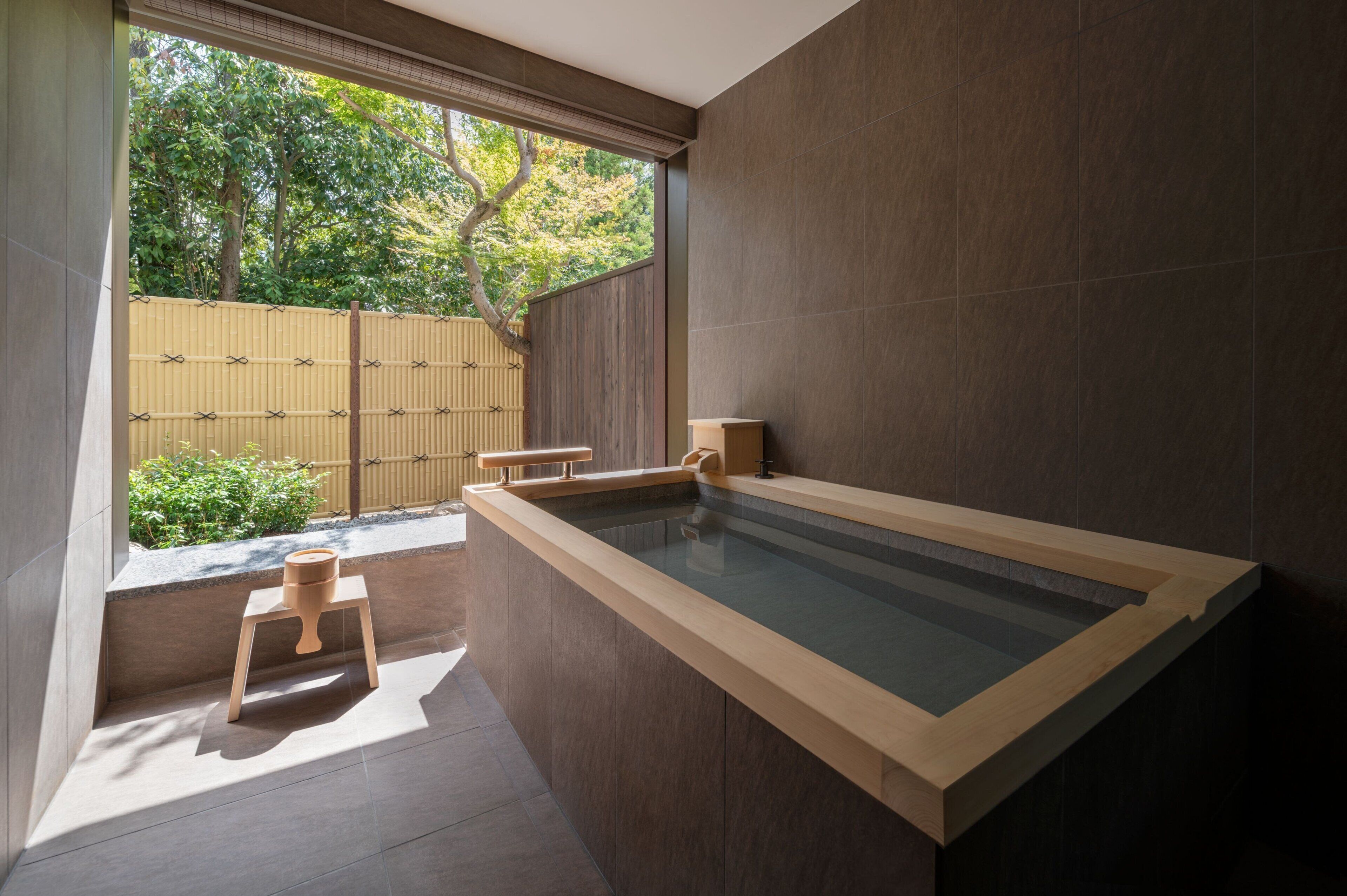 A modern bathroom with a large bathtub, a small wooden stool, and a bamboo fence visible through the window.