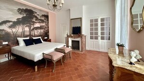 A bedroom with a large bed, a fireplace, a mirror, and a view of trees through the window.