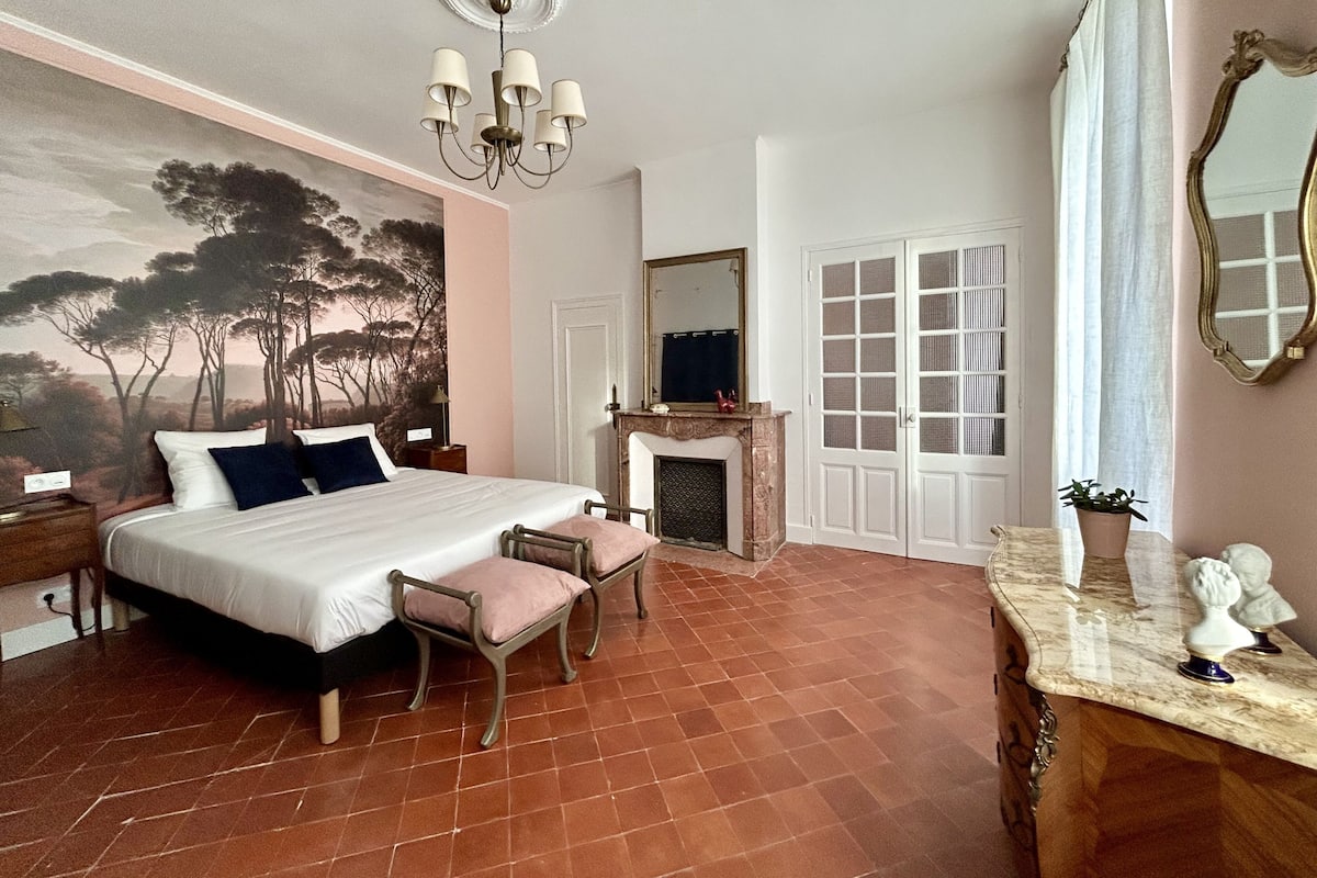 Un dormitorio con una cama grande, una chimenea, un espejo y vistas a árboles por la ventana.