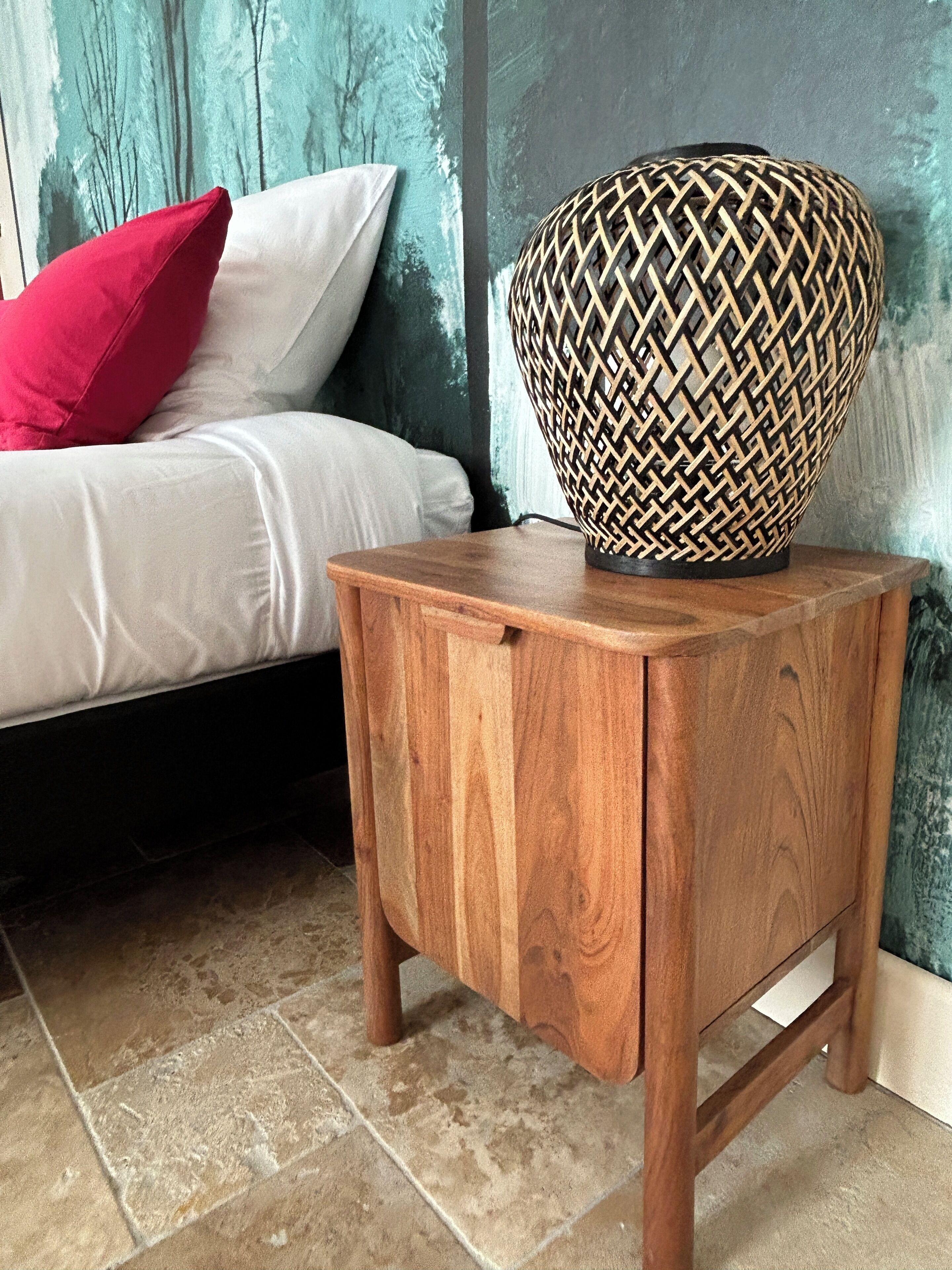 Una mesita de noche de madera con una lámpara de fibra trenzada junto a una cama con almohadillas blancas y rojas.
