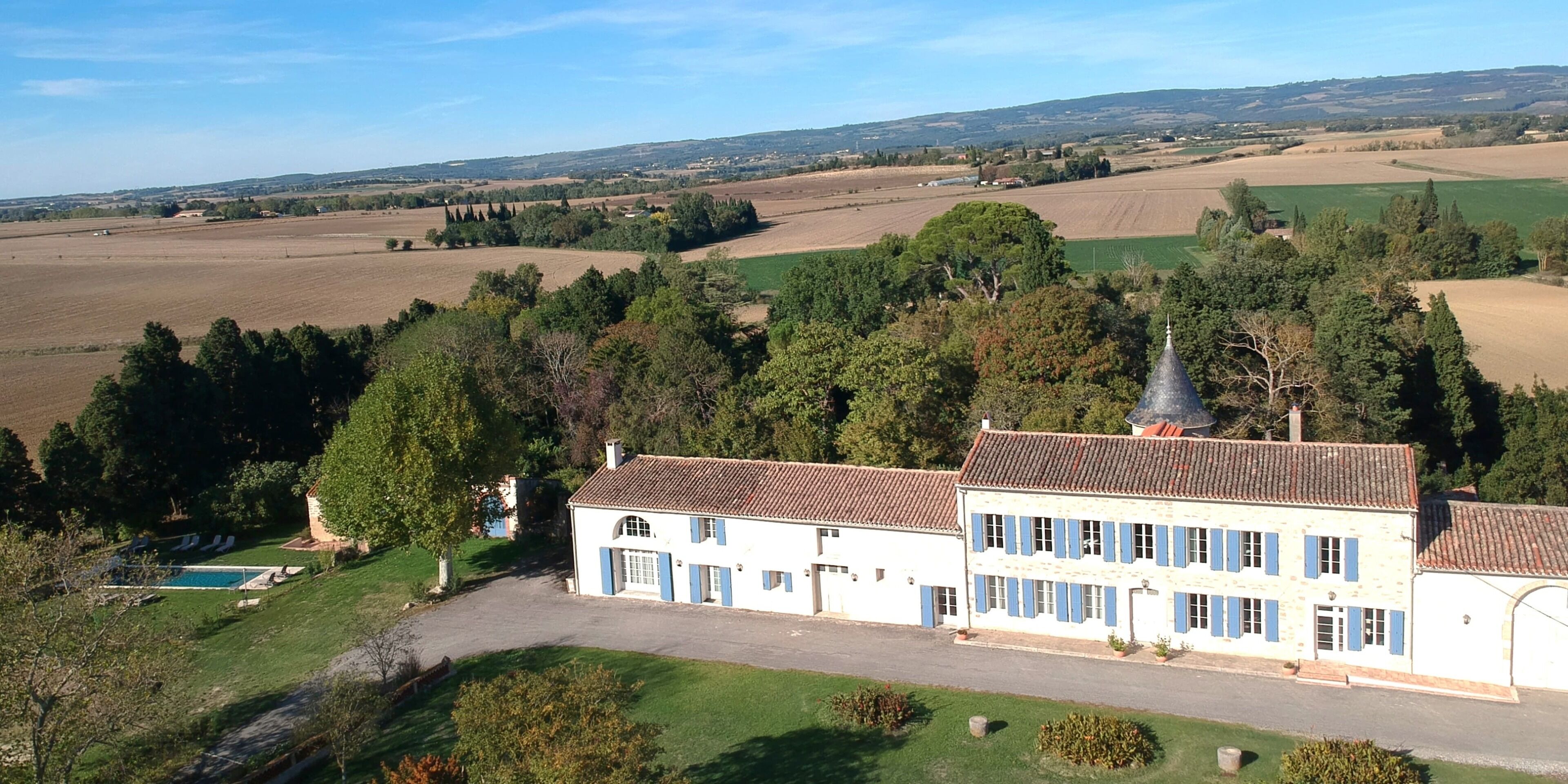 Un edificio grande de dos pisos, con techo de tejas y paredes blancas con contraventanas azules, rodeado de campos verdes y árboles.