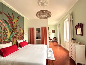 Un dormitorio con una cama grande, un aparador, una silla, una mesita, una estantería y un ventanal amplio.