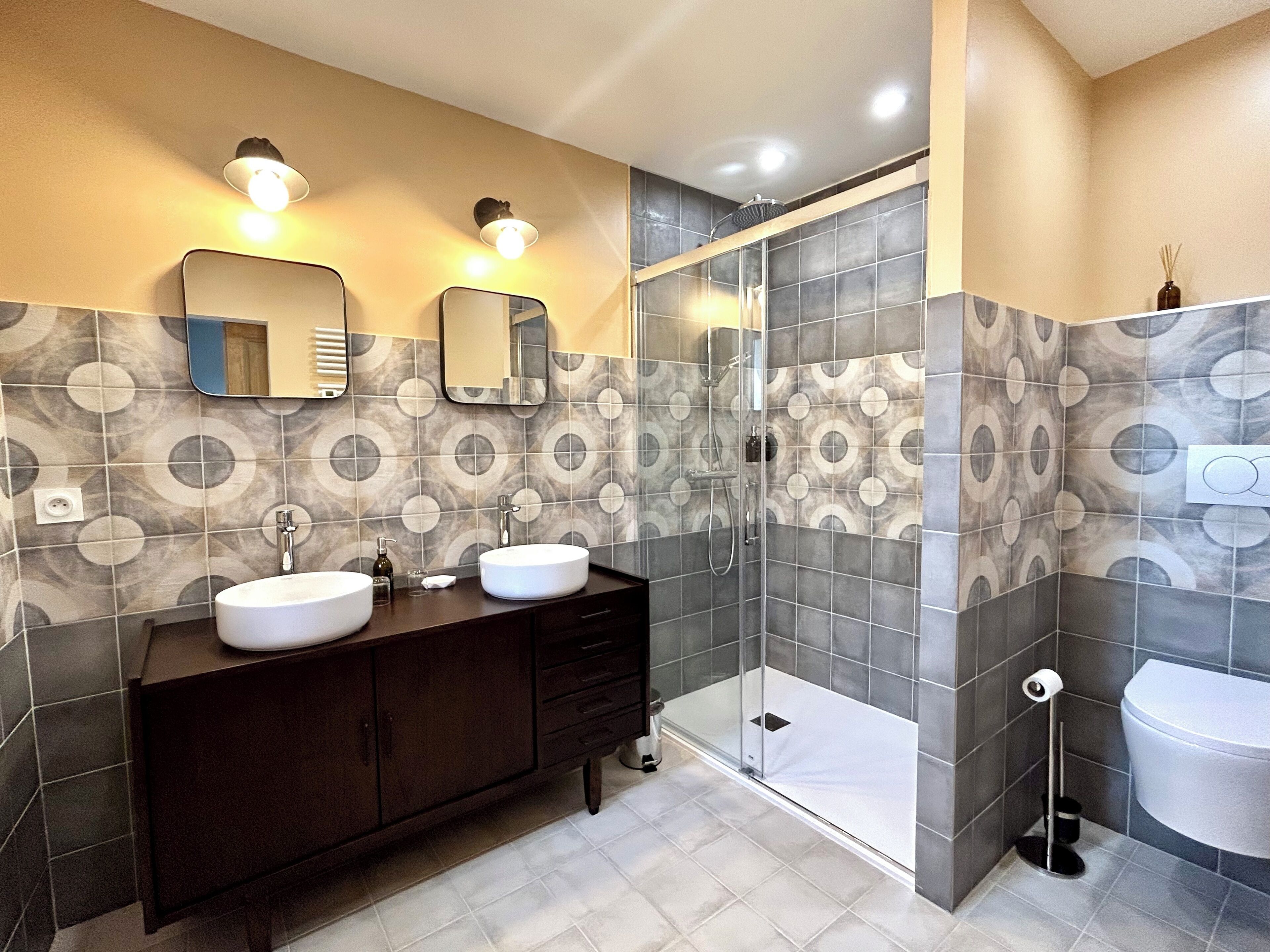 Une salle de bain avec deux lavabos, un meuble-lavabo foncé, une cabine de douche en verre et des carreaux à motifs.