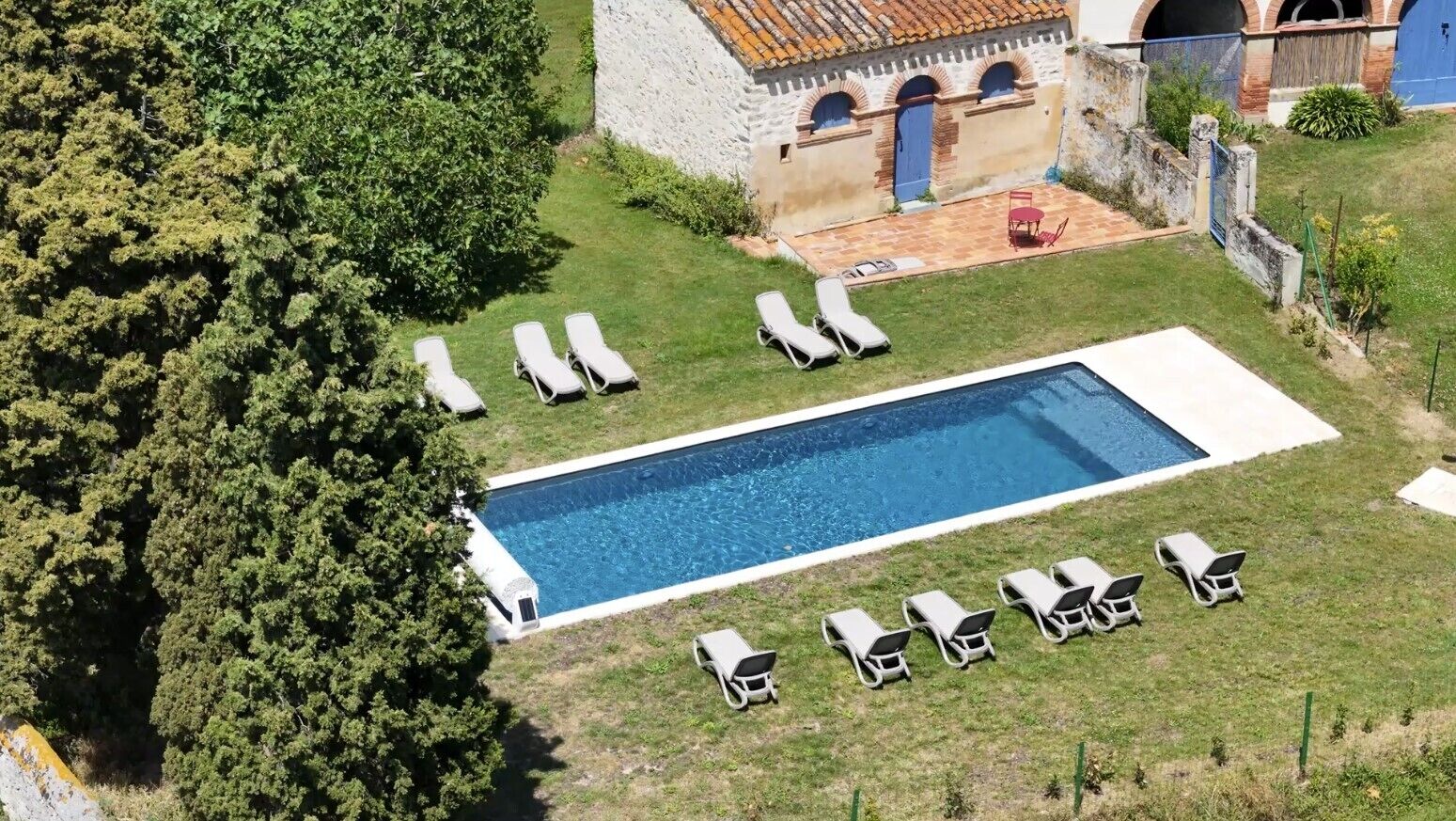 An aerial view of a property with a rectangular swimming pool surrounded by lounge chairs, a stone building with arches, and a garden area.