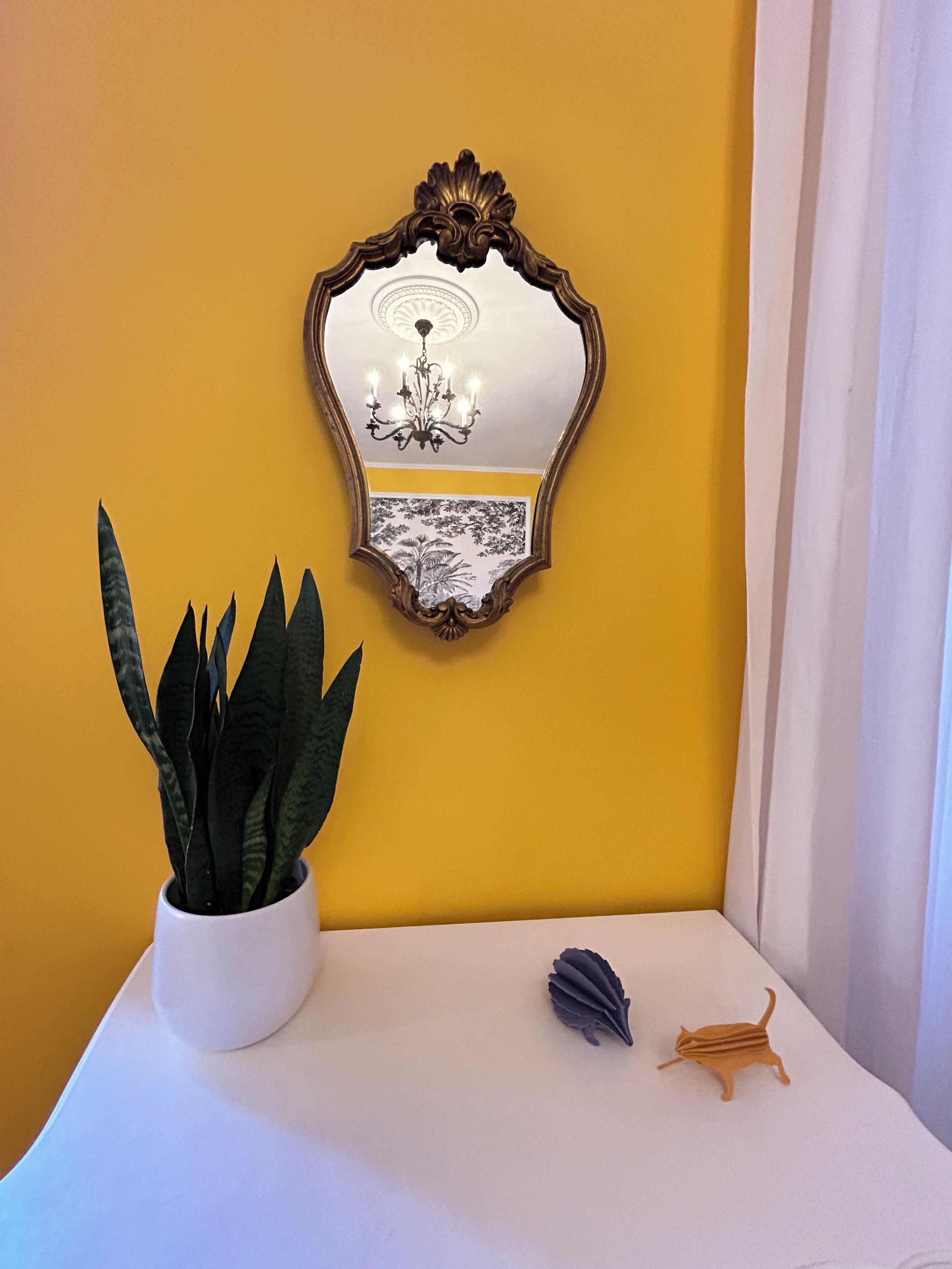 Una habitación con una pared amarilla, una mesa blanca, una planta en maceta, un espejo en marco y una pequeña figurita.