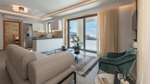 A modern living room with a sofa, a green armchair, and a dining area with a table and chairs. A kitchen is visible in the background with a large window. - Actea Seaview Villa II Free Heated Pool 800m sea (Kissamos)