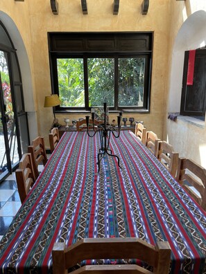 Un espace repas avec une longue table recouverte d’une nappe à motifs, entourée de chaises en bois, et une fenêtre donnant sur un paysage verdoyant.