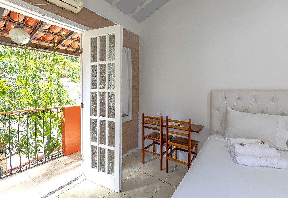 Un dormitorio con una cama, una silla de madera, una mesita y un balcón con vistas a la vegetación.