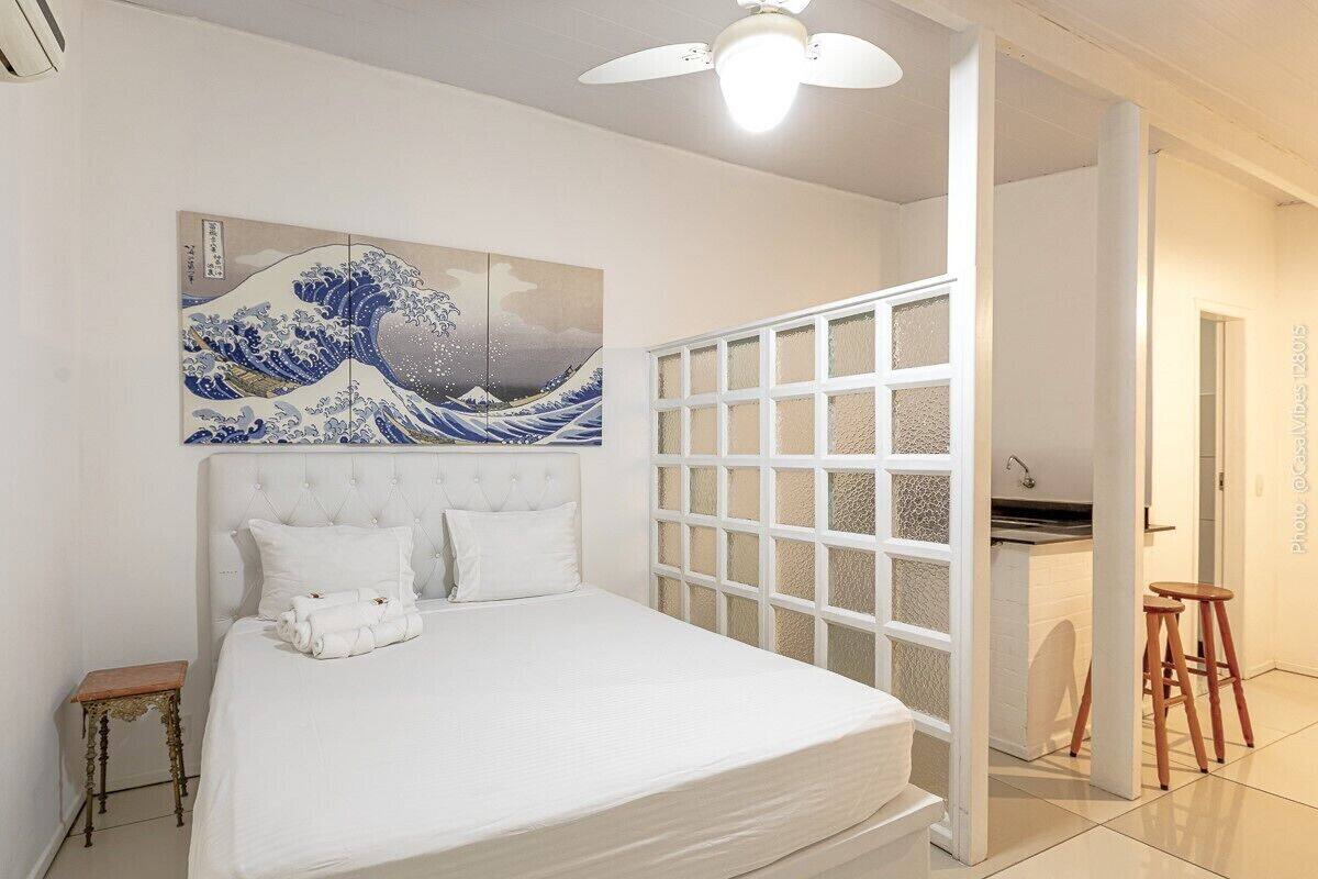 Une chambre à coucher avec un lit, une œuvre d’art accrochée au mur, une cloison blanche et une salle de bain visible à travers une porte ouverte.