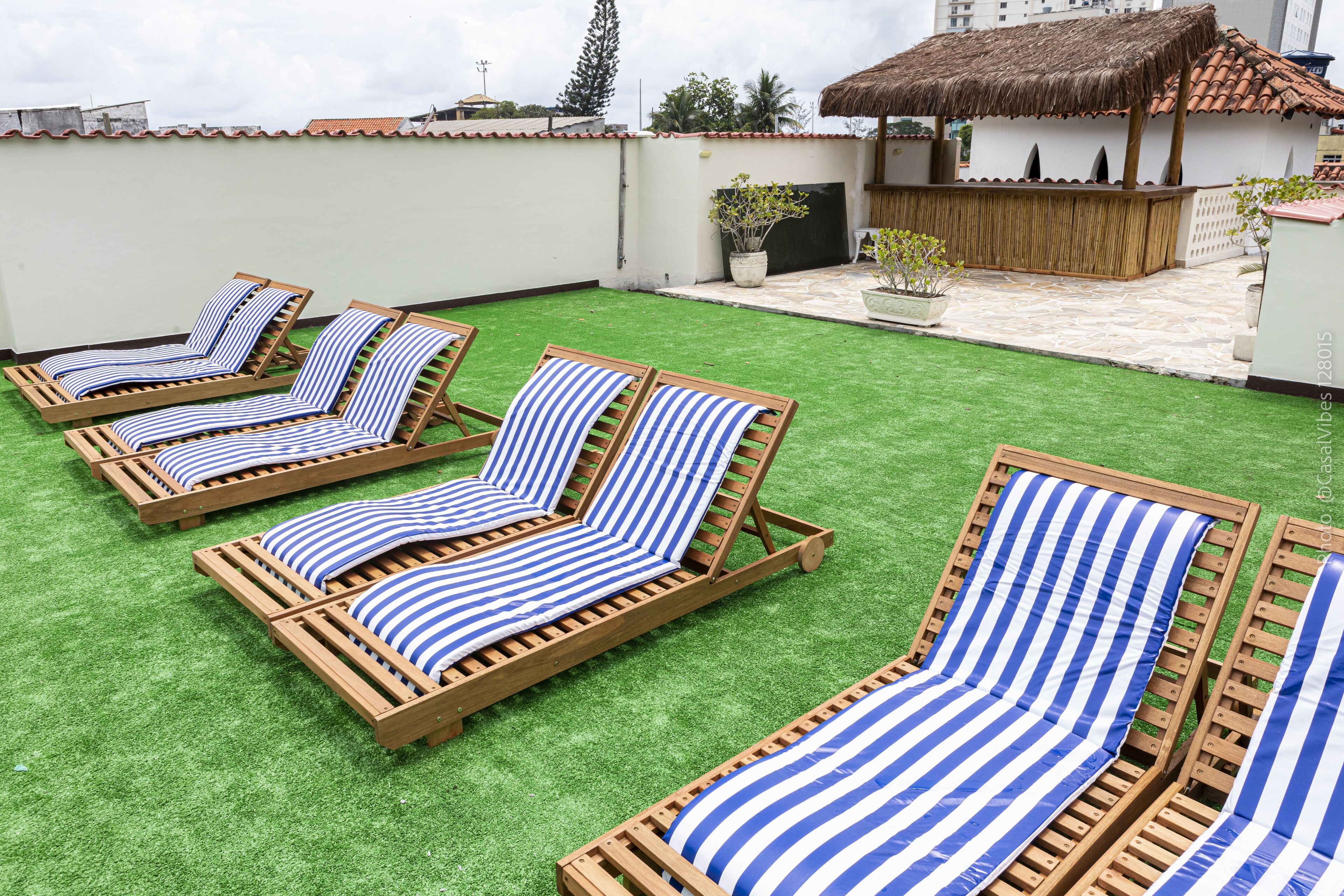 A rooftop with four wooden lounge chairs with blue and white striped cushions, a thatched-roof structure, and a potted plant.