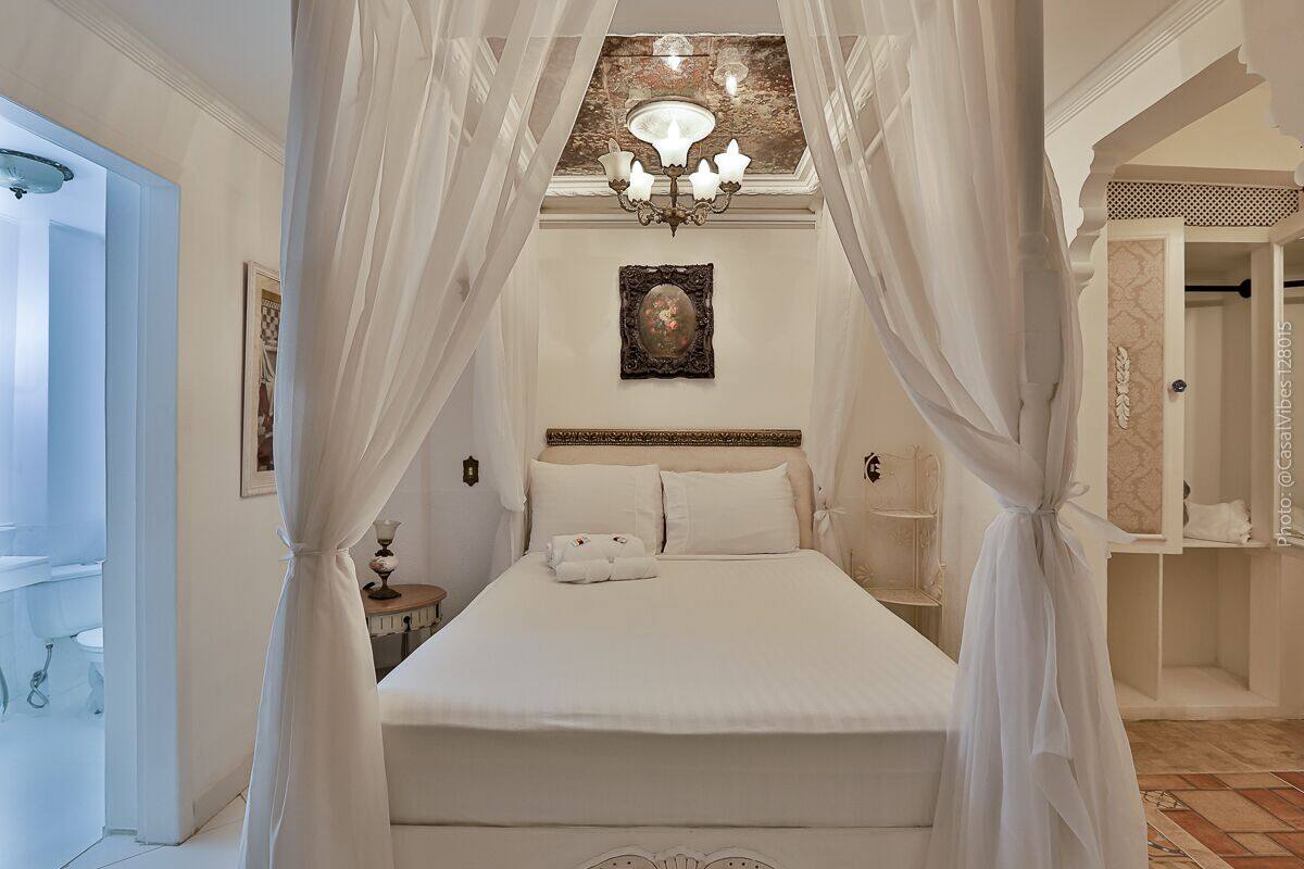 Une chambre à coucher avec un lit à baldaquin, un lustre, un tableau encadré au mur et une salle de bain visible à travers une porte ouverte.
