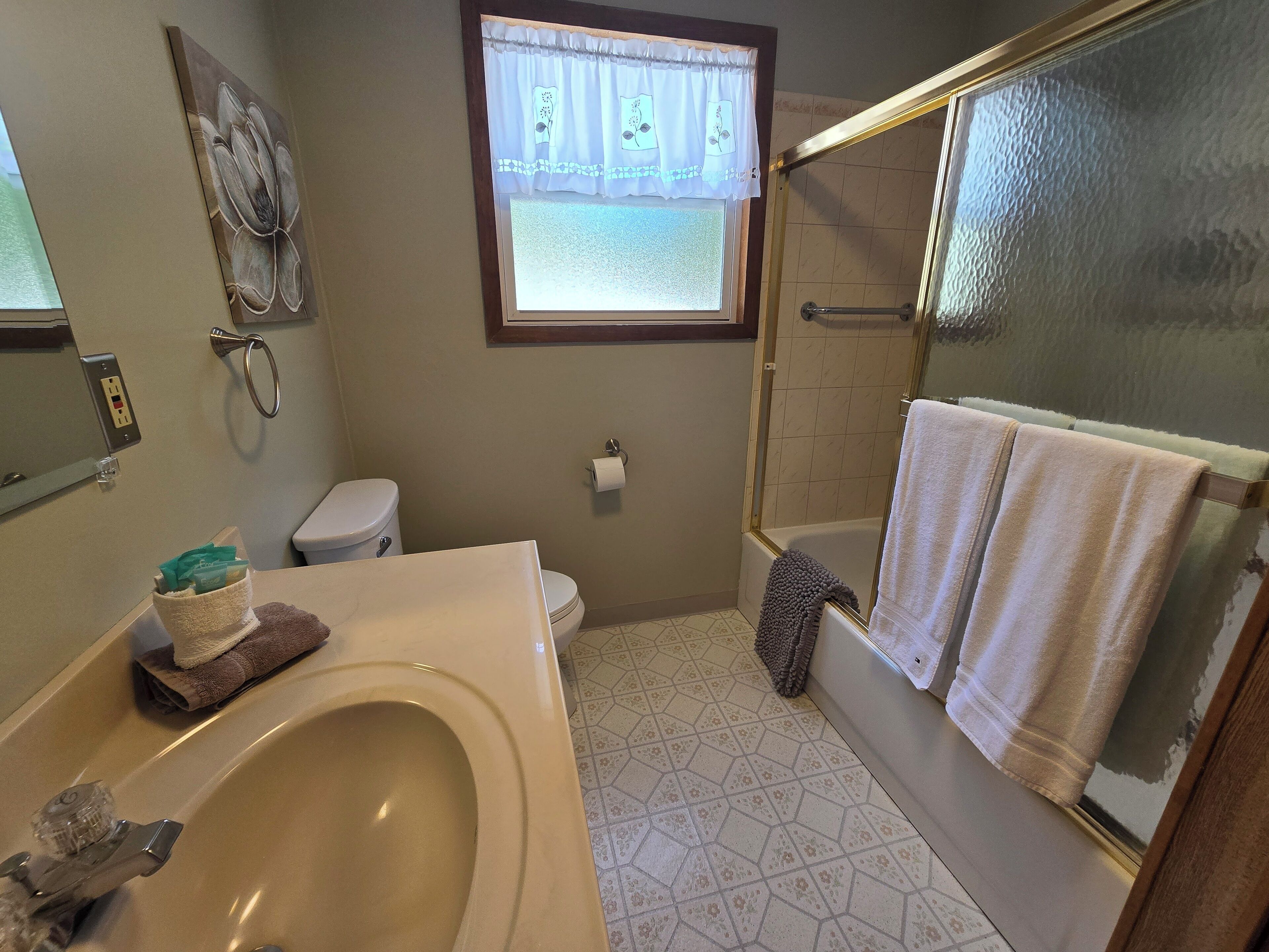 Ein Badezimmer mit Badewanne, Toilette und Waschbecken. Ein Fenster mit Vorhang, ein Handtuchhalter und eine Dusche sind vorhanden.