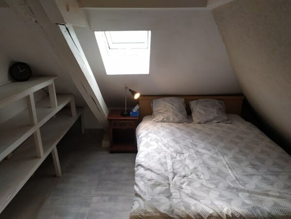 A small, sloped-roofed room with a single bed, a nightstand, a lamp, and a clock on a shelf.