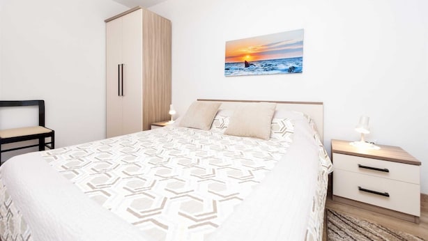 Une chambre à coucher comprenant un lit, une table de chevet, une armoire et un tableau représentant un coucher de soleil sur l’océan, accroché au mur.