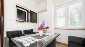 Une pièce avec une table à plateau en verre, des meubles en osier, un vase rempli de fleurs et une fenêtre avec des stores.
