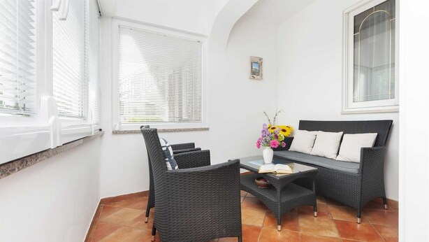 A room with a dark wicker sofa, two matching chairs, a small table with a vase of flowers, and a window with blinds. - MAXI One-Bedroom Apartment 2 (Rovinj)