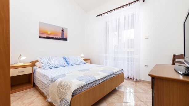 A bedroom with a bed, a nightstand, a desk, a lamp, a painting of a lighthouse, and a window with sheer curtains.