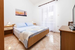 A bedroom with a bed, a nightstand, a desk, a lamp, a painting of a lighthouse, and a window with sheer curtains.