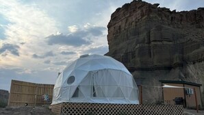 Ein kuppelförmiges Zelt mit Gitterbasis, vor einer Felsformation und einem Holzzaun gelegen.