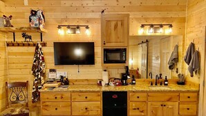 Eine gemütliche Blockhausküche mit Holzausstattung, einem schwarzen Kühlschrank, einer Mikrowelle, einem großen Flachbildfernseher und einer Arbeitsplatte aus Granit.