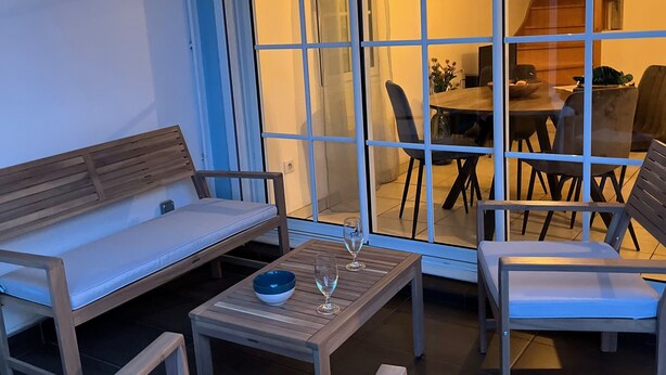 En utomhusplats med sittmöbler, bestående av en träbänk, två stolar och ett litet bord med en skål och vinglas.